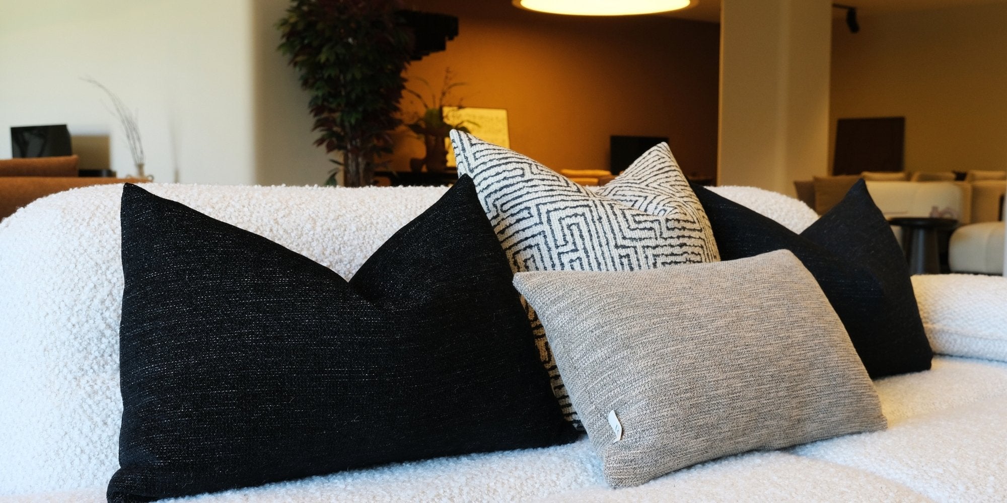 Remon Home dekoratif kırlent kombinasyonu; siyah dokulu kırlentler, geometrik desenli kırlent ve vizon tonlu kırlent ile krem buklet koltuk üzerinde modern, lüks salon dekorasyonu oluşturuyor.