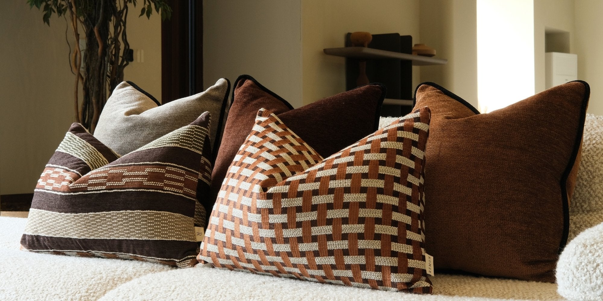 Remon Home dekoratif kırlent kombinasyonu; kahverengi düz, çizgili ve geometrik desenli premium kırlentlerle krem buklet koltuk üzerinde sıcak, modern ve lüks salon dekorasyonu oluşturuyor.